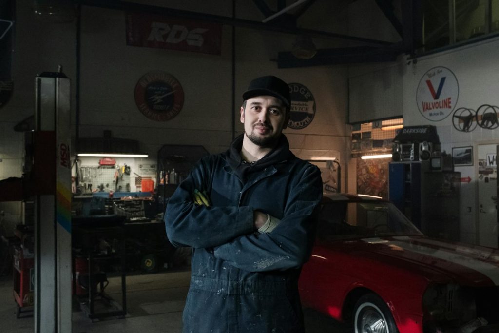 Experienced mechanic in a vintage garage standing confidently beside a classic car.