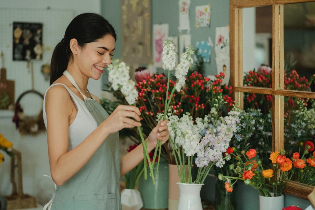 Side view of smiling florist with long dark hair in apron standing near counter and smelling flowers in vases in cozy floristry studio