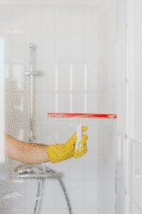 A person wearing a yellow glove cleans a shower glass with a squeegee, emphasizing hygiene and cleanliness.